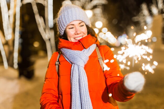 Belle jeune femme en bonnet et écharpe debout dans la ville avec la lumière du Bengale, cierge magique. Célébration du concept et Noël.