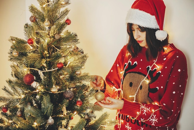 Belle jeune femme en bonnet de noel main tenant un ornement de paillettes dorées décorant le sapin de noël avec des lumières préparation pour les vacances dans un décor de salle de fête pour les vacances d'hiver