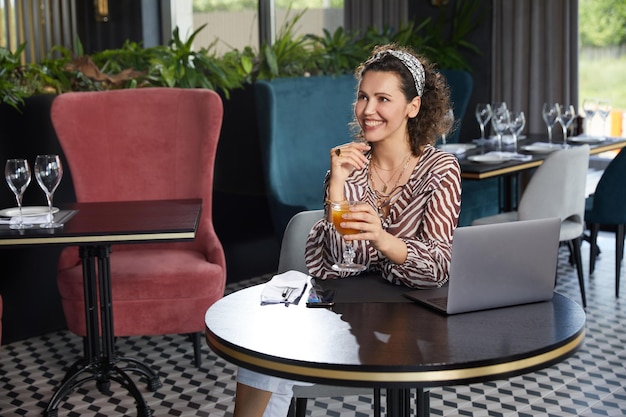 Belle jeune femme bouclée assise dans un café buvant du jus d'orange frais et attendant son amie