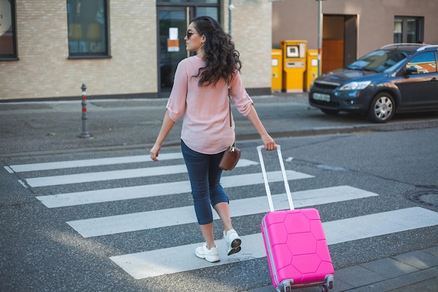 Belle jeune femme bouclée avec une petite valise rose est pressée de partir en vacances Un mannequin aux cheveux longs traverse la route dans une ville européenne Voyages
