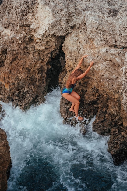 Une belle jeune femme bronzée aux cheveux longs en bikini bleu assis sur des rochers près de bleu vert clair