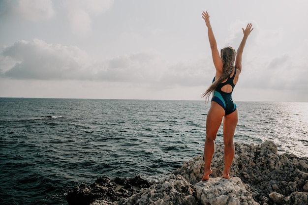 Une belle jeune femme bronzée aux cheveux longs en bikini bleu se tient au rocher près du bleu vert clair