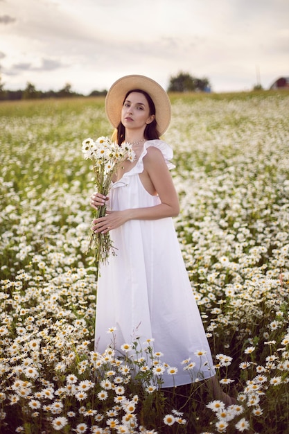 Belle jeune femme brune aux cheveux longs dans un chapeau et une robe blanche tient un bouquet dans ses mains debout sur un champ de camomille au coucher du soleil en été