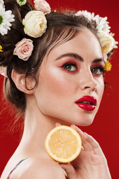 Belle jeune femme brune avec une coiffure florale incroyable portant du maquillage posant avec du citron isolé sur un mur rouge