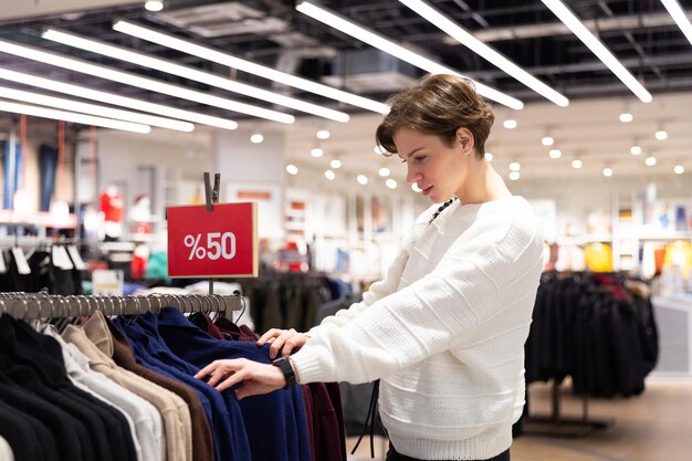 Belle jeune femme brune avec une coupe de cheveux courte dans un chandail blanc choisit des vêtements élégants dans un magasin dans un centre commercial