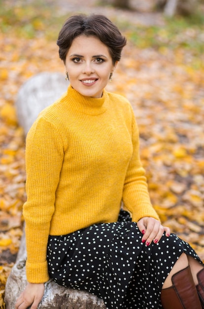 Belle jeune femme brune dans une jupe pull en tricot jaune et des bottes posant dans le parc d'automne et souriant