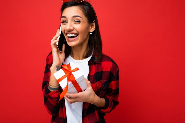 Belle jeune femme brune heureuse isolée sur un mur de fond coloré portant des vêtements décontractés élégants tenant une boîte-cadeau et regardant sur le côté.