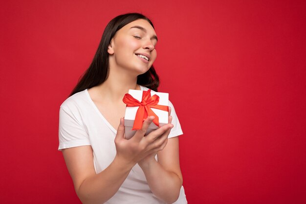 Belle jeune femme brune heureuse isolée sur un mur de fond coloré portant des vêtements décontractés élégants tenant une boîte-cadeau et regardant sur le côté.