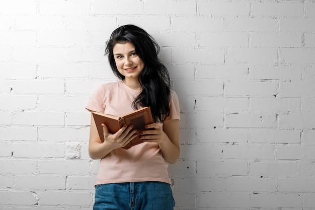 Belle jeune femme brune avec un livre debout près du mur de briques.