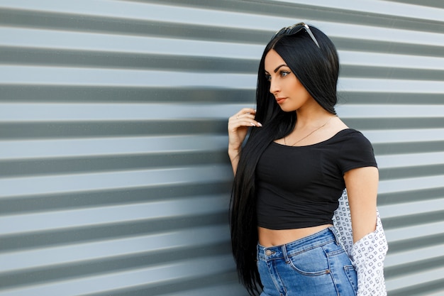 Belle jeune femme brune à la mode avec de longs cheveux noirs dans une cape élégante blanche en jeans vintage dans un t-shirt élégant noir près du mur moderne en métal avec des lignes dans la journée d'été