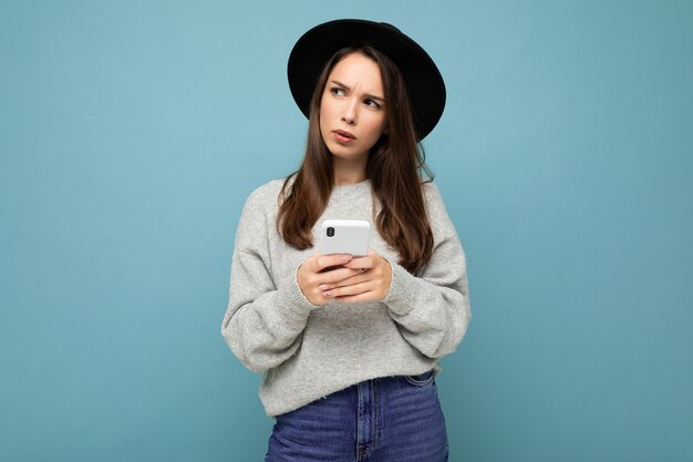 Belle jeune femme brune pensant portant chapeau noir et pull gris tenant le smartphone