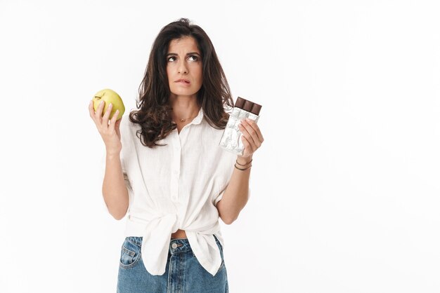 Belle jeune femme brune pensive portant des vêtements décontractés, isolée sur un mur blanc, tenant une assiette de chocolat et une pomme
