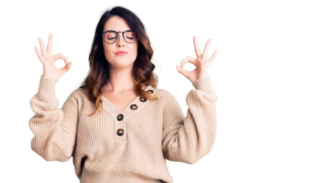 Belle jeune femme brune portant des vêtements décontractés et des lunettes détendue et souriante avec les yeux fermés faisant un geste de méditation avec les doigts concept de yoga