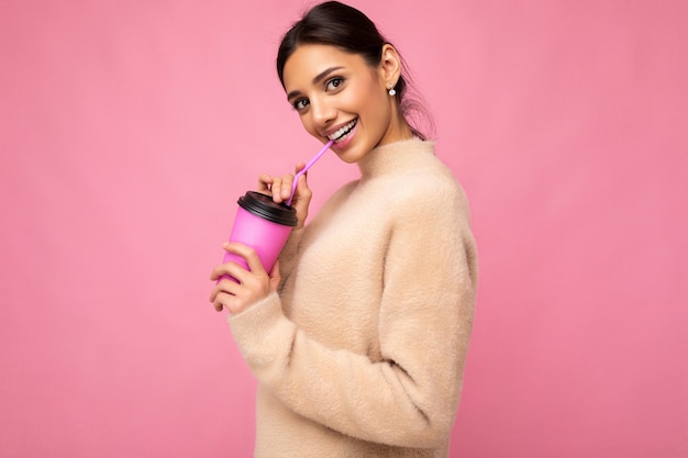 Belle jeune femme brune positive portant des vêtements élégants décontractés isolés sur un mur de fond coloré tenant une tasse de papier pour une maquette de boire du café en regardant la caméra.