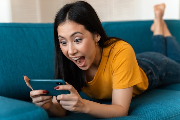 une belle jeune femme brune ravie émotionnellement surprise sur un canapé à l'intérieur à la maison à l'aide d'un téléphone portable.