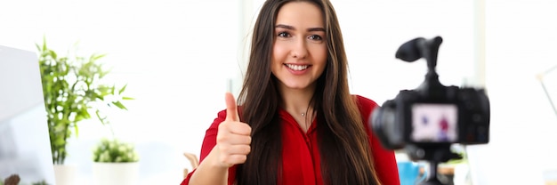 Belle jeune femme brune souriante en chemise rouge assis dans le bureau en face de la caméra sur trépied et montrant le pouce de pouce de bonne chance vers le haut.