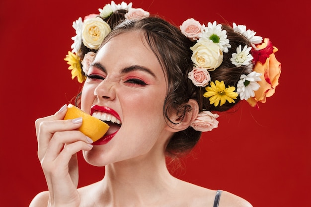 Belle jeune femme brune souriante avec une incroyable coiffure florale portant du maquillage posant isolée sur un mur rouge, mangeant du citron