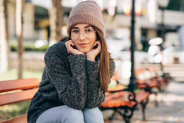 Belle jeune femme brune souriante largement portant des lunettes pull et chapeau en tricot Portrait en plein air d'un joli modèle féminin assis sur le banc dans la rue