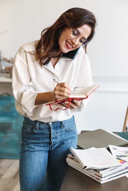 Belle jeune femme brune souriante parlant au téléphone portable tout en étudiant dans le café à l'intérieur, debout à la table