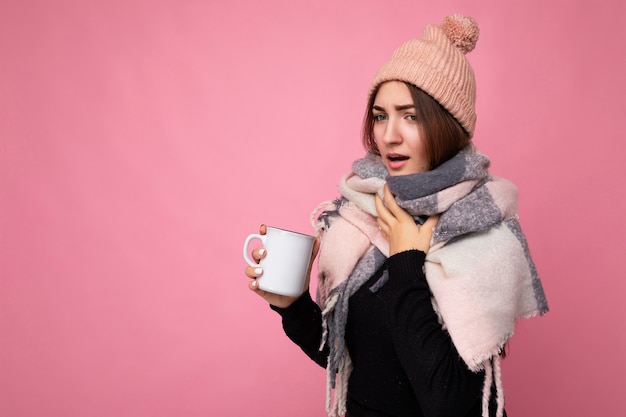 Photo belle jeune femme brune triste et malade portant un chapeau de chandail noir et une écharpe chaude isolée sur fond rose tenant une tasse en papier pour une maquette buvant du thé chaud et caughing. espace de copie