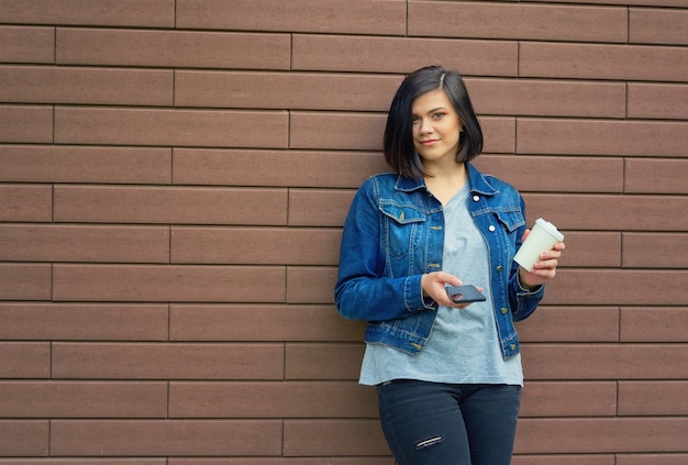 Belle jeune femme brune avec des tunnels dans les oreilles dans une veste en jean bleu avec une tasse de café et un smartphone debout devant le mur de briques.