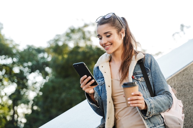 Belle jeune femme brune vêtue d'une veste, transportant un sac à dos marchant à l'extérieur, à l'aide de téléphone portable, tenant une tasse de café