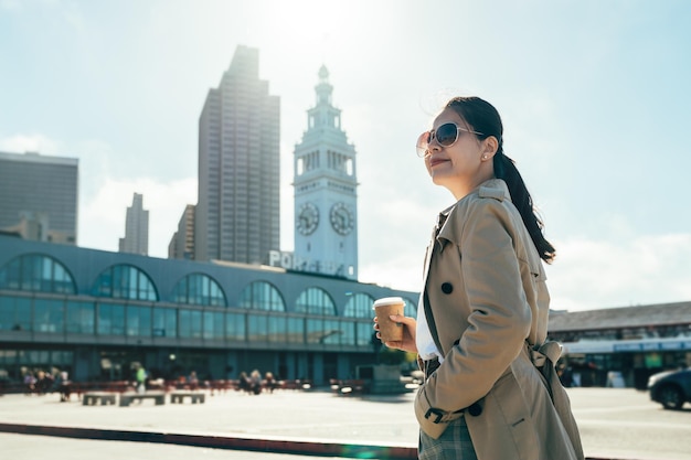 belle jeune femme de bureau tenant une tasse de café marchant sur la place devant le bâtiment du ferry à san francisco sous la lumière du soleil en été. femme d'affaires élégante se détendre profiter du tourisme.