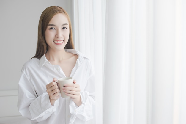 Belle jeune femme buvant du café debout près de la fenêtre de la maison.