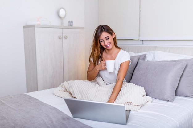 Belle jeune femme buvant du café à la maison dans son lit portant un t-shirt confortable tout en travaillant sur son ordinateur portable. Vérification des e-mails le matin, concept indépendant