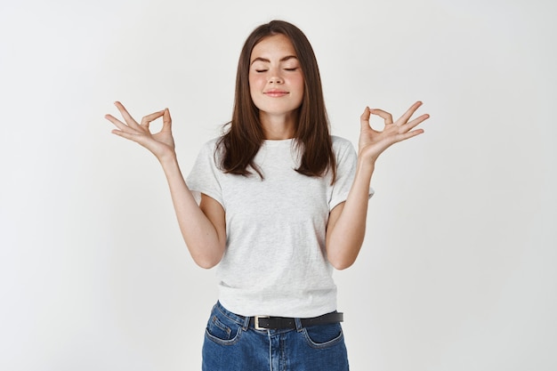 Belle jeune femme calme gardant les yeux fermés tout en pratiquant le yoga à l'intérieur, méditant, se tenant la main dans un geste de mudra, pensant à la paix