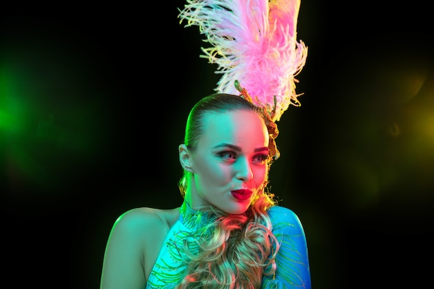 Belle jeune femme en carnaval, costume de mascarade élégant avec des plumes sur un mur noir à la lumière au néon