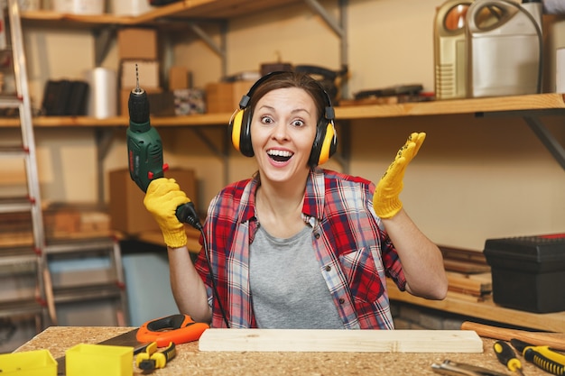 Belle jeune femme caucasienne aux cheveux bruns en chemise à carreaux, écouteurs insonorisés écartant les mains pour la joie, travaillant dans un atelier de menuiserie à table, perçant avec une perceuse électrique dans un morceau de bois.