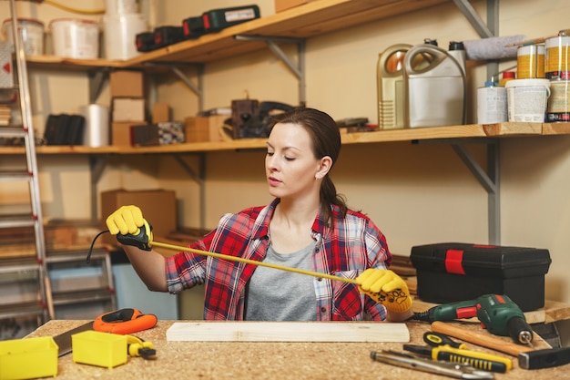 Belle jeune femme caucasienne aux cheveux bruns en chemise à carreaux, T-shirt gris, gants jaunes travaillant dans un atelier de menuiserie sur une table en bois avec différents outils, mesurant la longueur de la barre par un ruban à mesurer