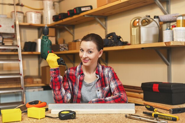 Belle Jeune Femme Caucasienne Aux Cheveux Bruns En Chemise à Carreaux Et T-shirt Gris Travaillant Dans Un Atelier De Menuiserie à Table, Perçant Des Trous Dans Des Morceaux De Fer Et De Bois Tout En Fabriquant Des Meubles.