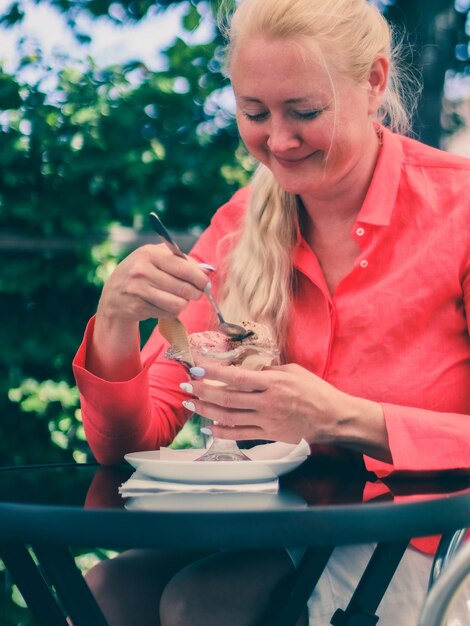 Une belle jeune femme caucasienne blonde tient un verre et mange de la crème glacée aux fraises au chocolat