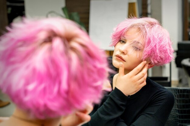 La belle jeune femme caucasienne avec une nouvelle coiffure rose courte regardant son reflet dans le miroir vérifiant la coiffure dans un salon de coiffure