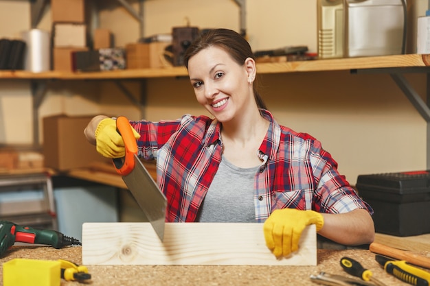 Belle jeune femme caucasienne souriante aux cheveux bruns en chemise à carreaux, T-shirt gris, gants jaunes sciant un morceau de bois avec une scie, travaillant dans un atelier de menuiserie sur une table en bois avec différents outils.