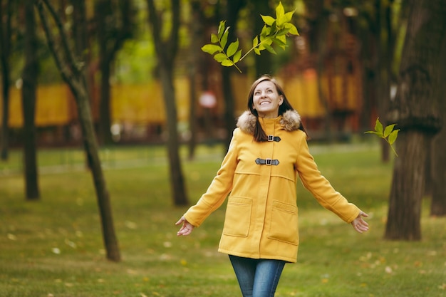 Belle jeune femme caucasienne souriante aux cheveux bruns en manteau jaune, jeans, bottes dans la forêt verte. Modèle féminin de mode vomir des feuilles d'automne debout et marcher dans le parc au début de l'automne à l'extérieur.