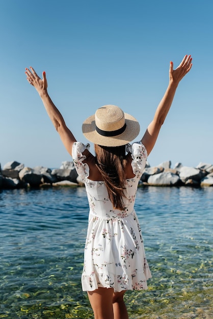 Une belle jeune femme avec un chapeau et une robe légère avec son dos se promène le long du rivage de l'océan sur fond d'énormes rochers par une journée ensoleillée Tourisme et voyages de vacances