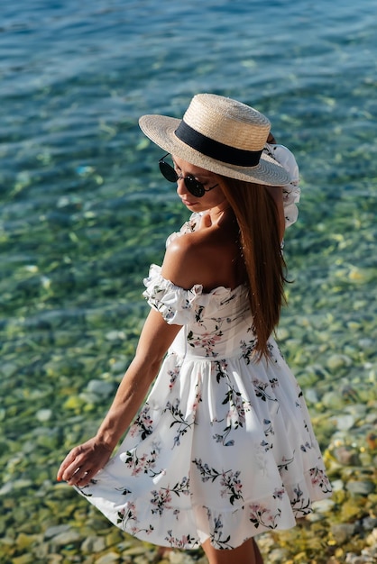 Une belle jeune femme avec un chapeau et une robe légère avec son dos se promène le long du rivage de l'océan sur fond d'énormes rochers par une journée ensoleillée Tourisme et voyages de vacances