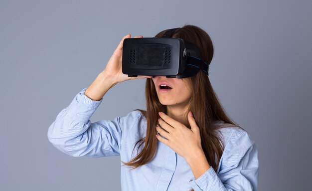 Belle jeune femme en chemise bleue à l'aide de lunettes VR et montrant la peur sur fond gris en studio