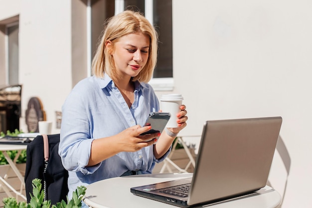 Belle jeune femme en chemise bleue travaillant sur un ordinateur portable dans un café et buvant du café Travail à distance