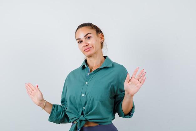 Belle jeune femme en chemise verte montrant un geste de reddition et l'air perplexe, vue de face.