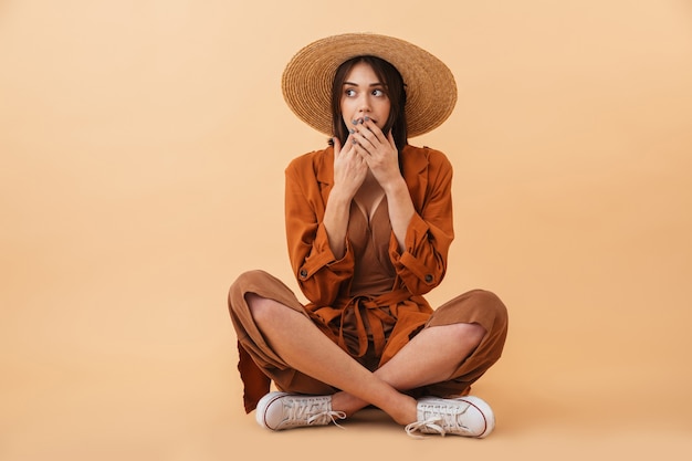 Belle Jeune Femme Choquée Portant Un Chapeau De Paille Et Une Tenue D'été Assise Isolée Sur Un Mur Beige