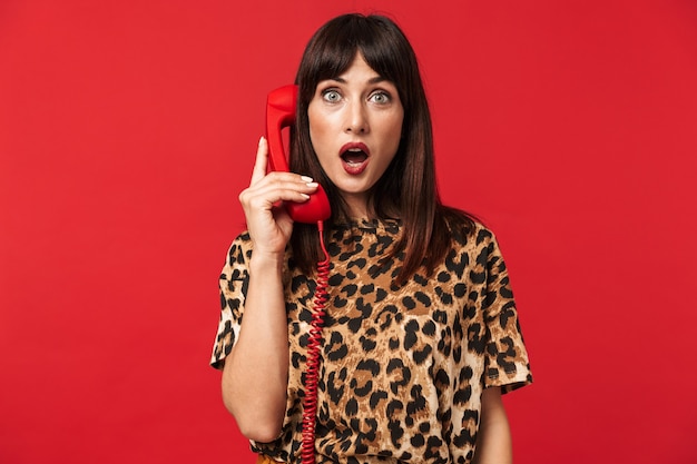 belle jeune femme choquée vêtue d'une chemise imprimée d'animaux posant isolée sur un mur rouge parlant par téléphone.