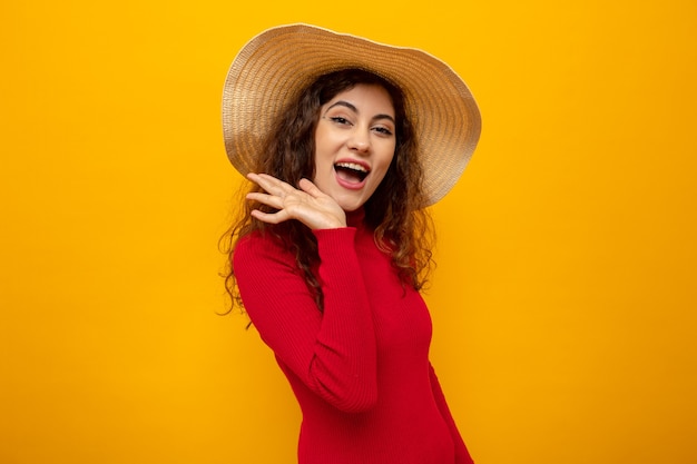 Belle jeune femme en col roulé rouge en chapeau d'été heureux et joyeux souriant largement debout sur orange