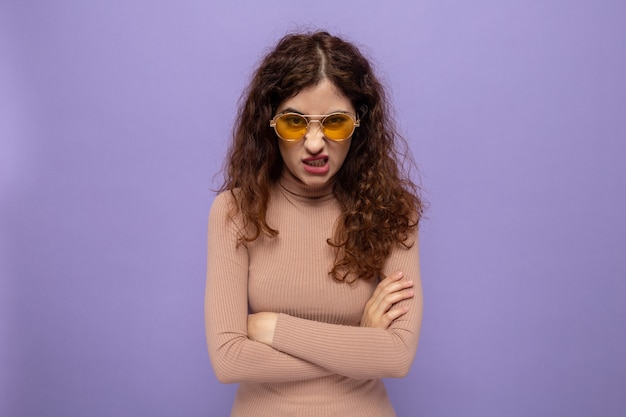 Belle jeune femme en colère en col roulé beige portant des lunettes jaunes regardant avec le visage fronçant les sourcils avec les bras croisés
