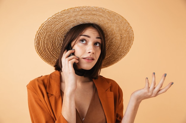 Belle jeune femme confuse portant un chapeau de paille et une tenue d'été isolée sur un mur beige