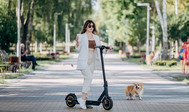 Belle jeune femme en costume blanc et lunettes de soleil debout avec son scooter électrique et Corgi Dog dans le parc de la ville et boire du café