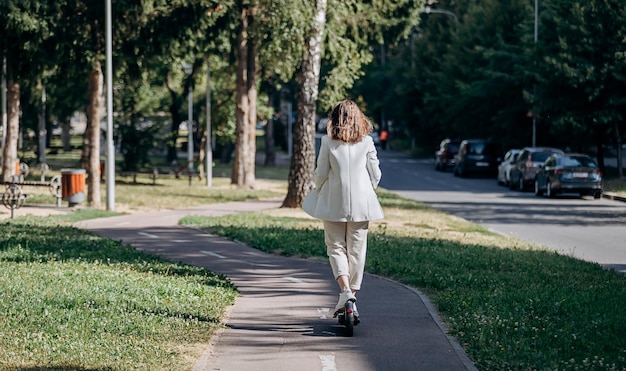 Belle jeune femme en costume blanc monte pour travailler sur son scooter électrique dans le parc de la ville